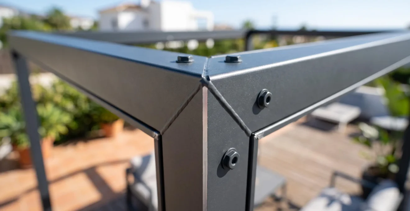 Plano detalle extremadamente cercano de la esquina de un perfil de aluminio de pérgola, con enfoque nítido en la textura del acabado lacado y profundidad de campo reducida que difumina el fondo de la terraza