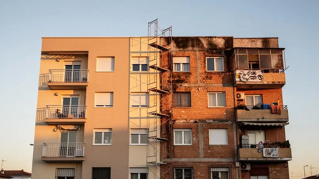 Edificio residencial español de los años 70 mostrando la mitad de su fachada ya rehabilitada con aislamiento térmico y la otra mitad aún sin intervenir, simbolizando la priorización de reformas con presupuesto limitado.