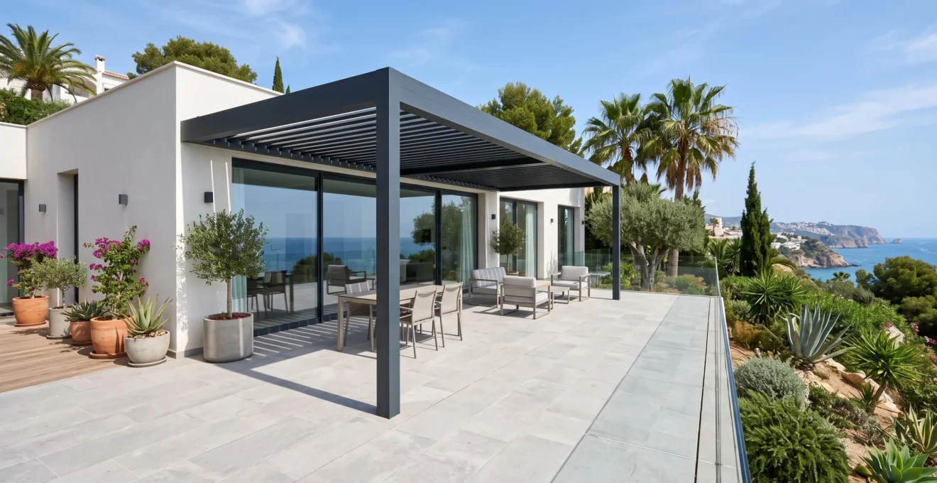 Vista de ángulo amplio de una terraza española contemporánea vacía con pérgola de aluminio adosada a una vivienda moderna, vegetación mediterránea de fondo con palmeras y olivos, cielo azul despejado, lamas de la pérgola abiertas y suelo de gres porcelánico imitación madera